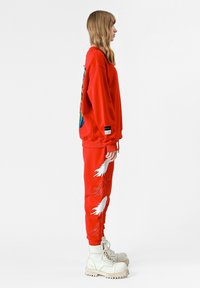 Sudadera roja con un estampado gráfico colorido, combinada con pantalones deportivos rojos que tienen diseños de plumas blancas, y botas gruesas blancas.