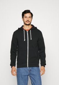 Zwart zip-up hoodie met witte trekkoordjes, geribde manchetten en een kangoeroezak, gedragen over een rode shirt, gecombineerd met blauwe spijkerbroek.