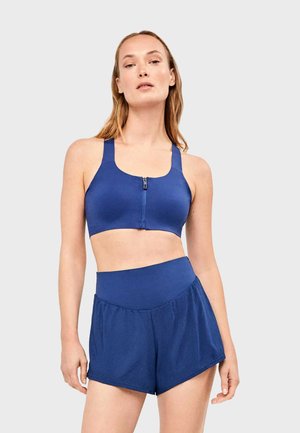 Vrouw draagt blauwe sportbeha met rits aan de voorkant en blauwe, hoog getailleerde mesh sportshorts, staand tegen een effen grijze achtergrond.