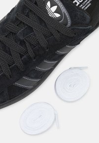 Černé semišové tenisky s černými tkaničkami, třemi bílými pruhy a kulatým černým logem. Vedle boty jsou položeny dvě bílé tkaničky.