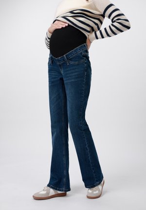Maternity jeans in dark blue denim, flared leg, featuring a stretchy black belly panel. Worn with light-colored casual shoes.