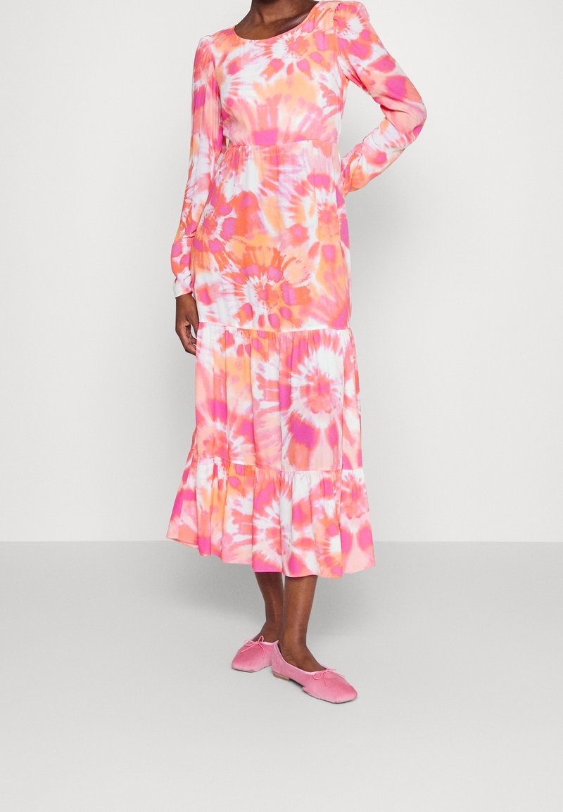 Femme portant une robe midi manches longues teinte en rose, orange et blanc avec une jupe à volants, associée à des ballerines roses.