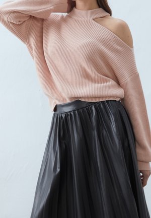 Femme portant un pull en maille rose clair avec une épaule dénudée et une jupe plissée noire taille haute.