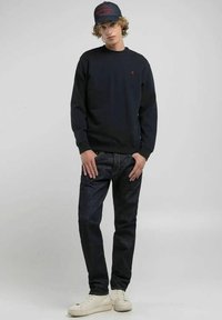 Felpa navy, collo a giro, logo rosso, jeans blu scuro, sneakers beige, cappellino nero con testo rosso, vestibilità rilassata, design minimalista.