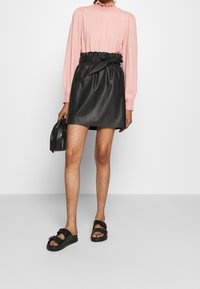 Gonna nera di pelle con cintura elastica e dettaglio a fiocco, abbinata a una blusa texturizzata rosa chiaro e sandali neri.