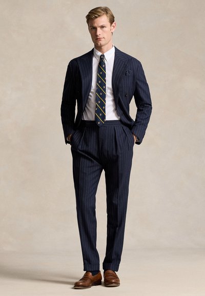 Mladý muž stojí s rukami v kapse, oblečený v tmavomodrom pruhovanom obleku, vzorovanej kravate, bielej košeli a hnedej koženej obuvi na béžovom pozadí.