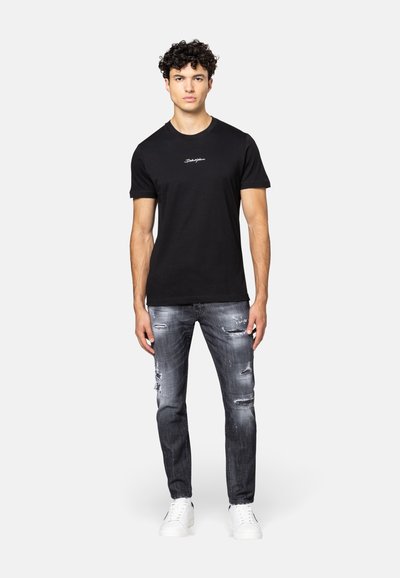 Zwarte katoenen T-shirt met korte mouwen, voorzien van een wit geborduurd logo; gecombineerd met versleten grijze jeans en witte sneakers.