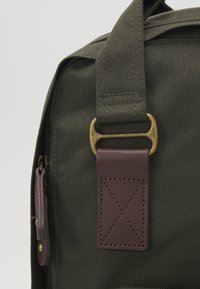 Gros plan sur le coin d'un sac en toile vert foncé avec une sangle en cuir marron passant à travers une boucle en laiton et une tirette de fermeture éclair en cuir.