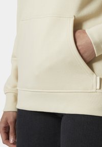 Crème hoodie met een voorzak, ribbelmanchetten en -zoom, met een zachte textuur en eenvoudig ontwerp, gedragen met een donkere spijkerbroek.
