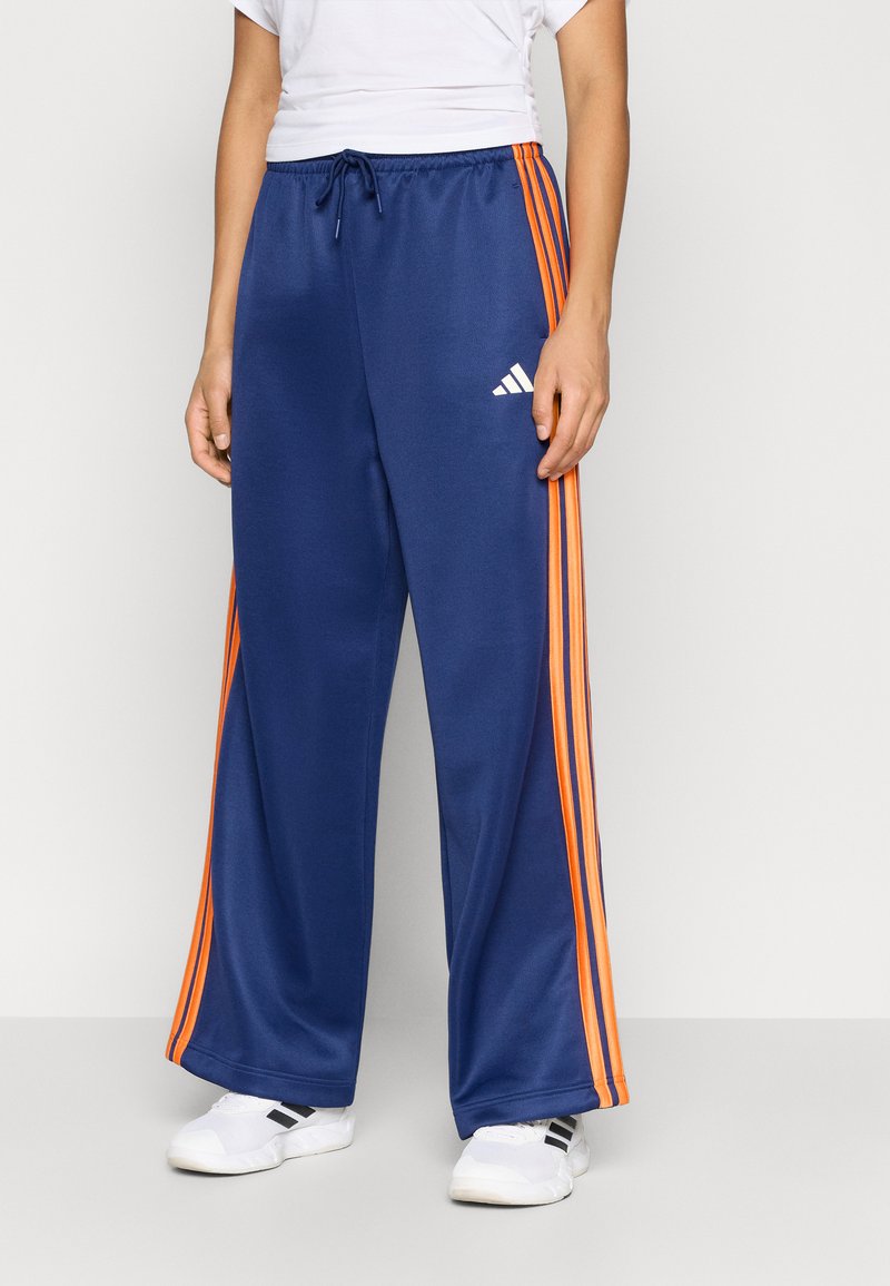 Blå treningsbukser med oransje stripete detaljer, laget av glatt stoff, med elastisk midjebånd og Adidas-logo.
