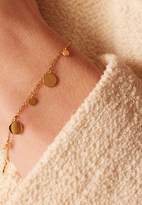 Gouden armband met kleine en middelgrote ronde bedels, bevestigd aan een delicate ketting, rustend op een gestructureerd huidoppervlak.