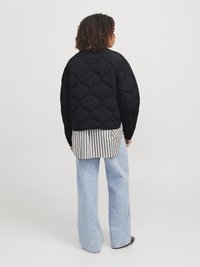 Personne ayant des cheveux bouclés portant une veste matelassée noire sur une chemise rayée et un pantalon large en jean bleu clair, se tenant devant un fond uni.