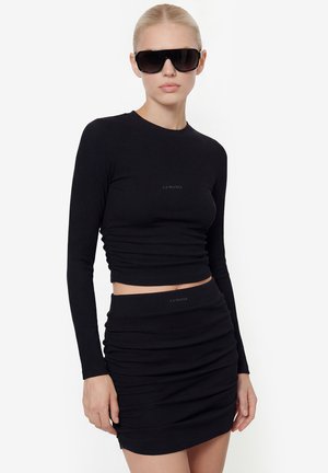 Žena nosi elegantný čierny dlhý crop top s dlhým rukávom a zladenú mini sukňu s naškrobenými detailmi, s čiernymi veľkými slnečnými okuliarmi, pózujúc na bielom pozadí.