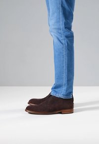 Ruskeat mokkanahkaiset nilkkurit, joissa on teksturoitu pinta, nauhakiinnitys ja puukanta, yhdistetty vaaleansinisiin istuviin farkkuihin.