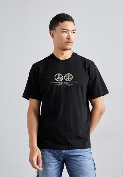 Zwarte katoenen T-shirt met korte mouwen, voorzien van drie cirkelvormige vredessymbolen in groen en beige, vergezeld van tekst in een eenvoudig lettertype.
