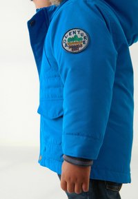 Lyst blå pufferjakke med foer i hætten, frontlomme og et cirkulært mærke med "ADVENTURE" på venstre arm.