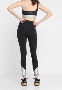 Černé sportovní legíny s lesklými stříbrnými akcenty, síťovými panely a přiléhavým designem, doplněné o černou sportovní podprsenku a zlaté tenisky.