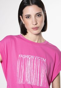 Lyserød t-shirt med korte ærmer, prydet med teksten "FASHION VICTIM" i hvid og frynse-detaljer under teksten. Glat tekstur.