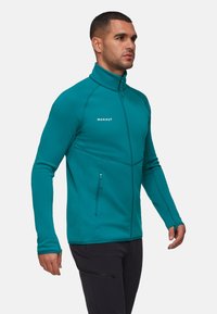 Teal fleece zip-up jack met hoge kraag, contrasterende naden en een zijvak met rits. Rekbare stof ontworpen voor mobiliteit en comfort.