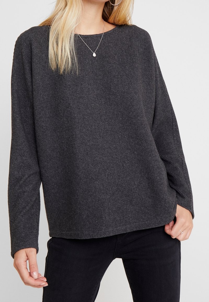 Szary sweter o luźnym kroju, żebrowanej fakturze i szerokim dekolcie. Ma długie rękawy i prosty dół, zestawiony z czarnymi spodniami.