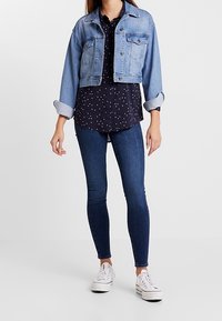Donna che indossa una giacca di jeans leggera e corta di colore blu chiaro su una camicia nera a fantasia, jeans skinny scuri e sneakers bianche, in piedi su uno sfondo bianco.