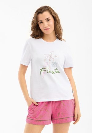 Femme aux cheveux bruns portant un t-shirt blanc « Fiesta » avec des motifs de palmiers et de perroquet et un short rose à motifs, les mains dans les poches.