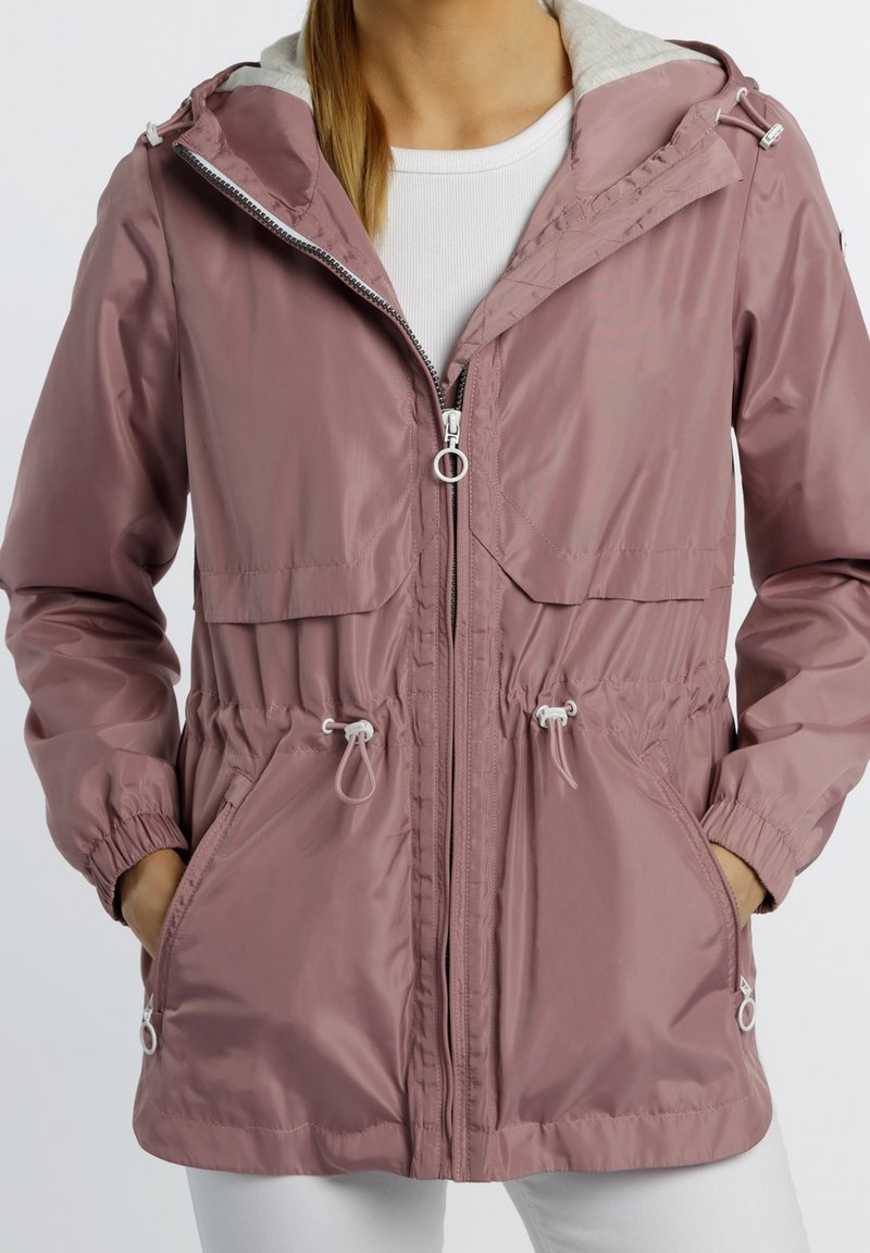 Femme portant une veste à capuche rose poudré avec fermeture éclair à l'avant, cordon à la taille, poignets élastiques et poches, par-dessus un haut blanc et un pantalon blanc.