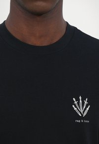 Camiseta de algodón negra con cuello redondo y un gráfico bordado en blanco de cinco dagas en el lado izquierdo del pecho.