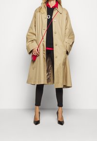 Manteau trench beige avec une coupe ample, un grand col et une fermeture à boutons. Porté par-dessus des leggings noirs et un sweatshirt avec des accents rouges, complété par un petit sac rouge.