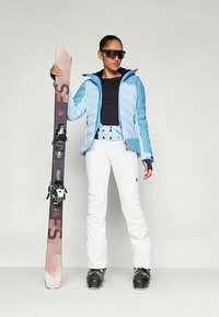 Σαλοπέτα σκι με ελαφρύ μπλε φουσκωτό μπουφάν, μαύρη μπλούζα, λευκά παντελόνια και μαύρες μπότες σκι. Κρατάει καστανά και μαύρα σκι με δέστρες.