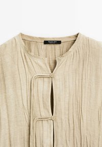 Haut en lin beige avec une finition texturée, doté d'un col rond, de coutures verticales et d'une fermeture à un bouton à l'avant.