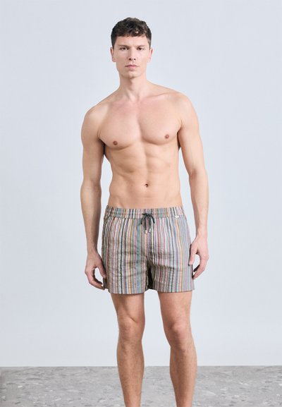 Νέος άντρας χωρίς πουκάμισο που στέκεται, φορώντας πολύχρωμα μαγιό με κάθετες ρίγες και κορδόνι, σε ουδέτερο φόντο.