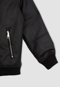 Chaqueta de cuero negra con un acabado texturizado, bolsillo con cremallera y puños acanalados. El diseño incluye costuras pronunciadas y un ajuste relajado.