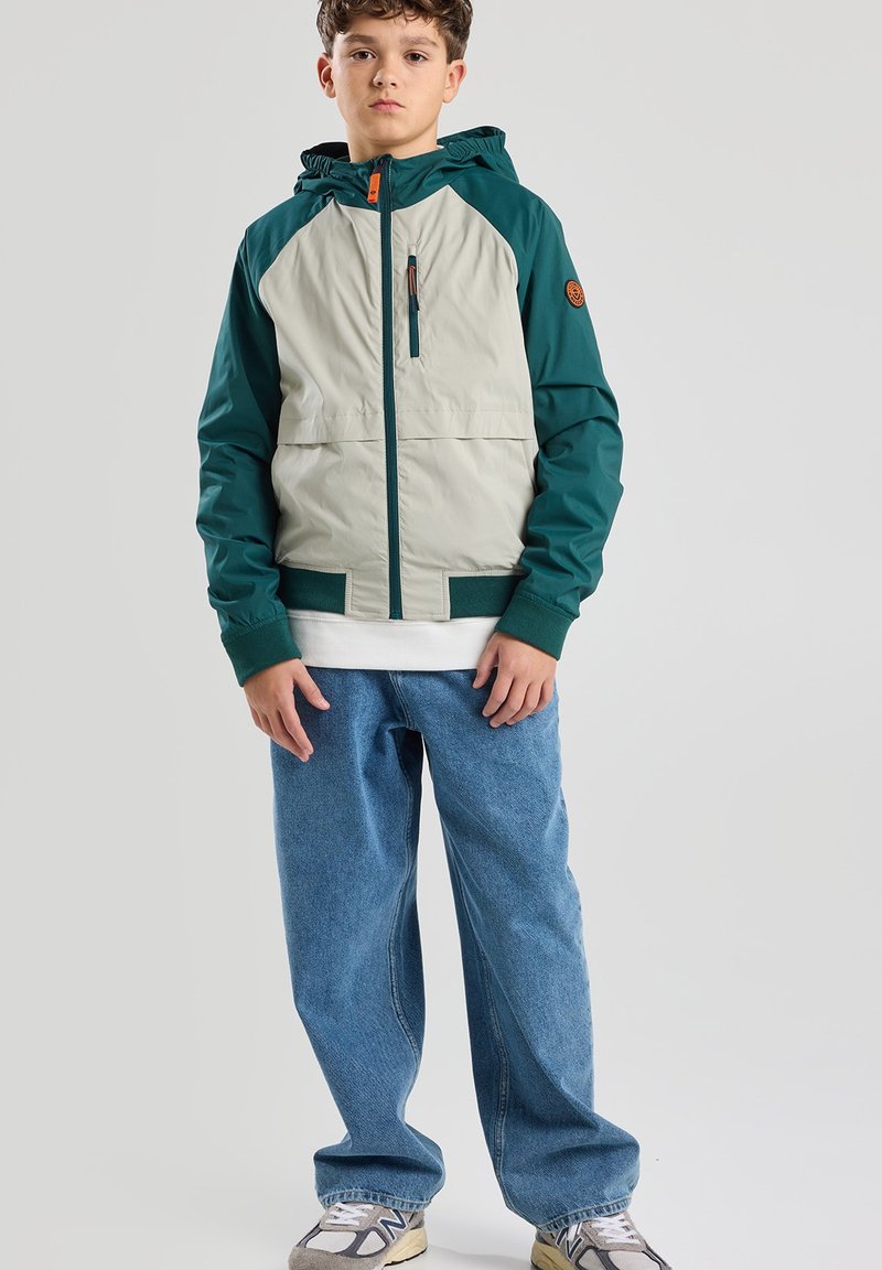 Giacca con cappuccio di colore verde scuro e grigio chiaro, dotata di una tasca con zip e polsini elastici, abbinata a jeans blu larghi e sneakers beige.