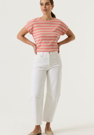 Vrouw draagt een roze-beige gestreept T-shirt met opgerolde mouwen, witte high-waist brede broekspijpen en beige platte schoenen, staand met handen in de zij.