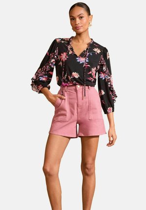 Sort blomsterbluse med lange ærmer, V-hals og rynkede manchetter; parret med lyserøde shorts med forlommer og en tætsiddende talje.