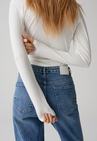 Weißes Langarmoberteil in Kombination mit hoch taillierten blauen Jeans. Das Hemd hat schmale Ärmel, und die Jeans verfügen über klassische Gesäßtaschen.