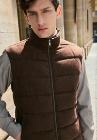 Brun puffer vest med lodret syning, høj krave og frontlynlås. Båret over en grå langærmet skjorte. Glat tekstur, afslappet design.
