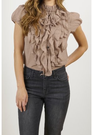 Beige Bluse mit geraffter Vorderseite und Tupfenmuster, kurzen Ärmeln und hohem Kragen, kombiniert mit dunkelgrauen Jeans.