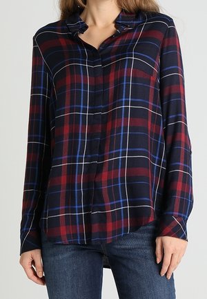 Kvinde iført en skjorte med lange ærmer og knapper med rød, blå og hvid ternet mønster, kombineret med blå jeans.