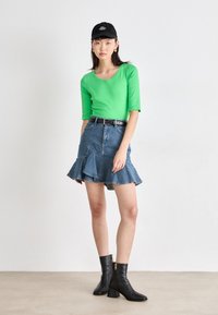 Haut vert ajusté, jupe en denim évasée avec un ourlet volan, ceinture noire et bottines noires à fermeture éclair. La mannequin porte une casquette noire.