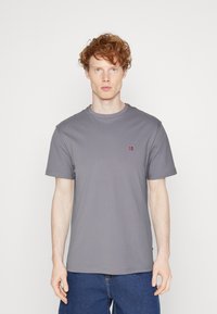 Grå crewneck t-skjorte laget av en bomullsblanding, med korte ermer og et lite rødt og blått emblem på brystet. Jevn tekstur.