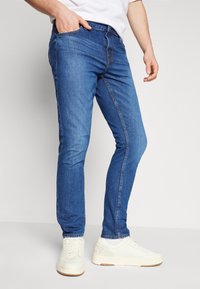 Slim-fit sinimärgid denim teksad, millel on kulunud tekstuur, viie taskuga disain, nupuga ja tõmblukuga kinnitamine, koos valgete spordijalatsitega.