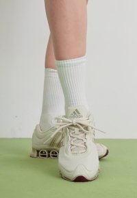Hvite sportsko med beige detaljer, mesh-øvre del og gummisåle. Kombinert med ribbede hvite sokker, plassert mot et grønt gulv.