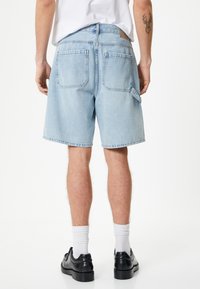 Lysblå denim shorts med løs pasform, der har baglommer og et mærke på taljen, båret med sorte sko og hvide sokker.