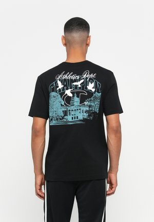 Mann trägt ein schwarzes T-Shirt mit blauer Grafik eines Gebäudes, Bäumen, weißen Vögeln und dem Text "Athletics Dept. Champion" auf dem Rücken.