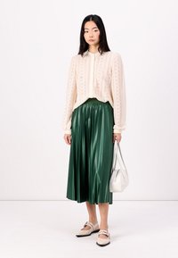 Cárdigan de punto crema, falda midi plisada verde, zapatillas beige y un bolso blanco. Texturas suaves, patrones ligeros y formas fluidas.