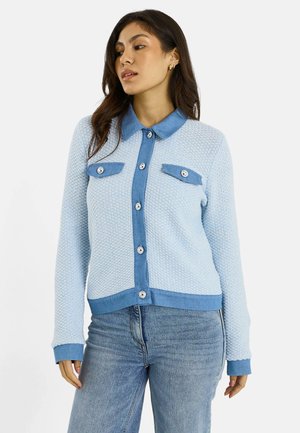 Mujer que lleva un cárdigan abotonado de textura azul claro con cuello, puños y tapetas de bolsillo de mezclilla, combinado con unos vaqueros azul claro.