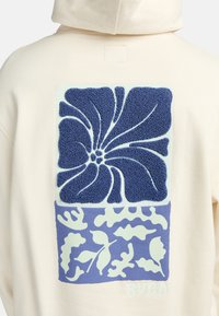 Sweat à capuche crème avec un graphique floral texturé bleu dans le dos. Présente des motifs en relief et un plus petit design de feuille en dessous. Le texte indique "RVCA."