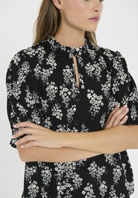 Blusa nera a fiori con motivi bianchi, colletto arricciato e maniche corte a sbuffo. Il tessuto ha un aspetto testurizzato. Dettaglio con apertura a chiave.